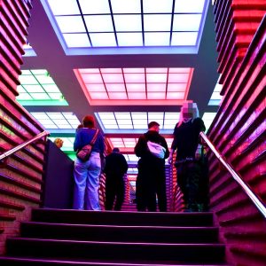 Menschen stehen oben an einer Treppe in einem Raum mit farbig beleuchteten Deckenfenstern und roten Ziegelwänden