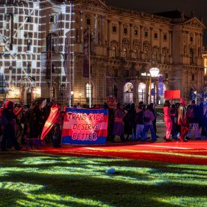 Demo for the Trans Day of Rememberance 2025, the lights on Heroes Square create interesting patterns on the lawn. In the middle of the demo train with a flag of TNYV.