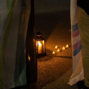 Demo zum Trans Day of Rememberance 2025, in der Mitte des Bildes sind Kerzen zu sehen. Durch die dunklen Lichtverhältnisse am Abend leuchten diese stark. Links und Rechts ist das Bild von gehaltenen Bannern abgeschnitten.