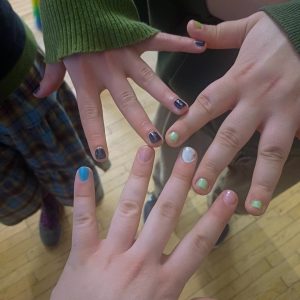 You can see three hands of three different people with spread fingers with the nail polish that they painted during the meeting. Background of the photo are the lower bodies of the three people and the wood floor in the room of the WUK where the meeting took place.