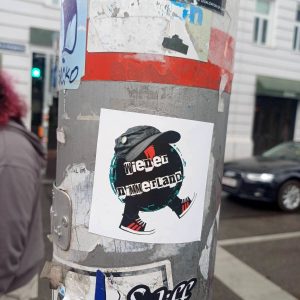 Im Mittelpunkt und nahe an der Kamera ist ein Sticker mit dem Logo von Wiener Nimmerland, das auf einer Straßenschildstange klebt. Rechts im Hintergrund ist unscharf ein blaue Limousine zu Links eine Person mit pinken Haaren zu sehen. Die Stange hat einige, teilweise abgekratzte, andere Sticker kleben. Die Stange ist silbern/metallisch und hat einen roten Streifen, so wie quasi alle Straßenschildstangen in Wien.