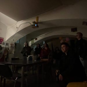 Dark room with several people, one person stands at a table leaning forward left with microphone, others standing or sitting in the background, table and chairs visible