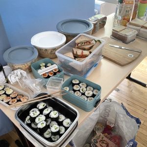 Auf einem Tisch stehen Behälter mit Sushirollen, Sandwiches und Salaten. Die Sushirollen sind in mehreren rechteckigen und runden Dosen, einige mit Sesam bestreut, andere mit Avocado oder Gurke gefüllt. Daneben sind Sandwiches mit unterschiedlichen Belägen in transparenten Boxen, ein Schneidebrett mit weiteren Sandwiches und ein Glas Wasser. Eine Flasche Sojasauce steht am Rand. Im Hintergrund sind weitere Lebensmittel und Getränke zu sehen.