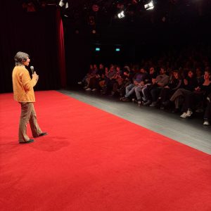 Person mit Mikrofon steht auf rotem Bühnenboden vor Publikum in dunklem Saal