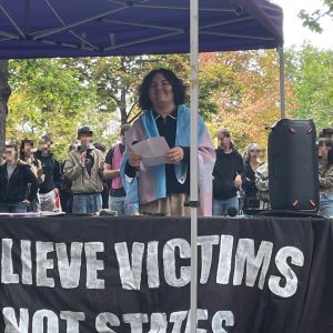 Person steht unter einem lila Pavillon und hält Papiere mit Notizen zur Rede, die die Person gerade hält, vor einem Banner mit der Aufschrift 'BELIEVE VICTIMS NOT STATES'. Um die Schultern trägt die Person eine trans Flagge.